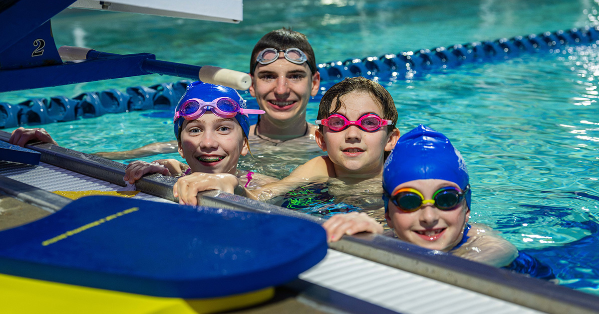 Family of four kids standing in a pool, smiling at the camera. They are all wearing goggles and two are wearing blue swimcaps.
