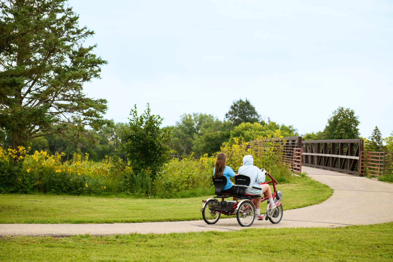 two people riding on an adaptive bike facing away from the camera on a bike trail