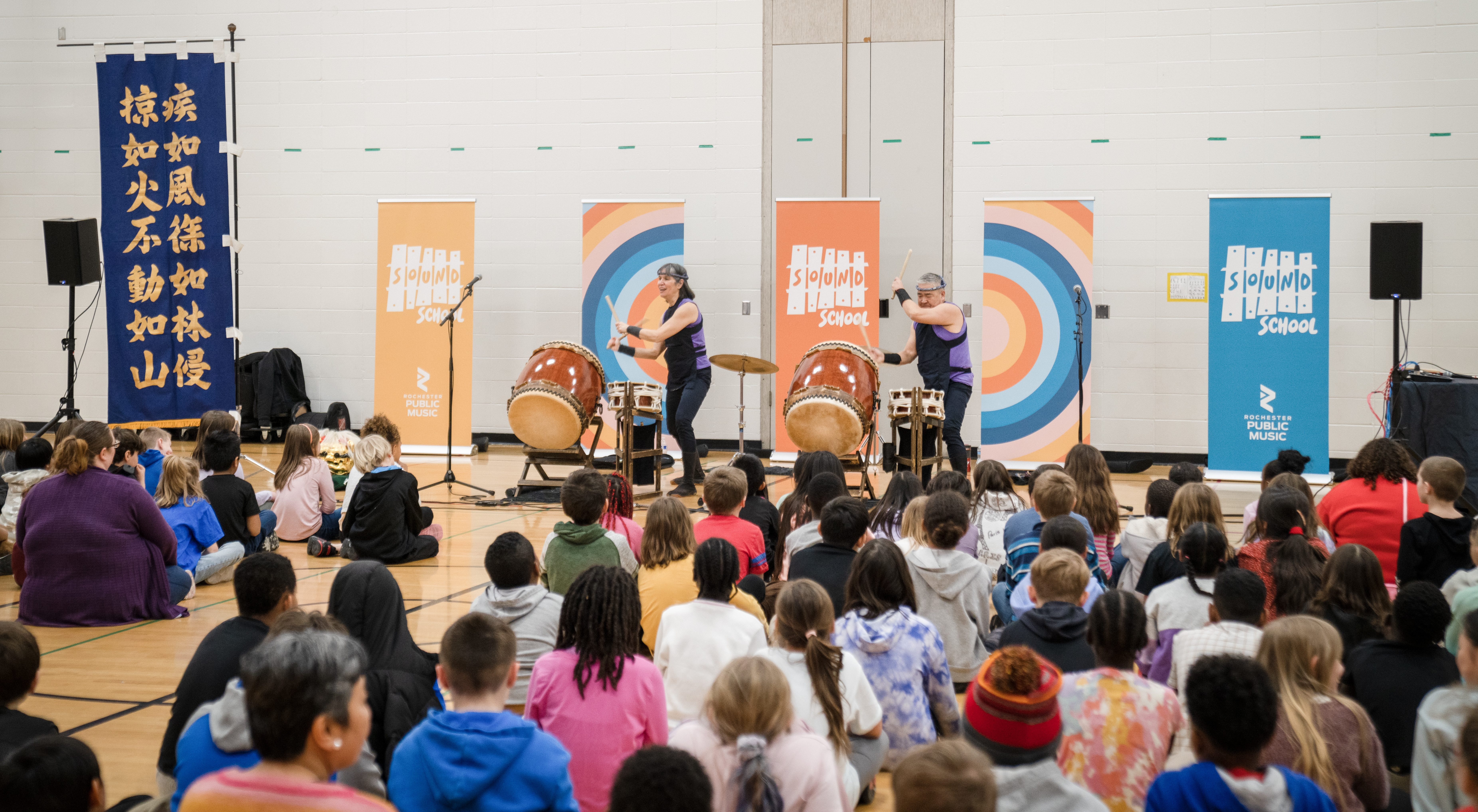 Students seated in a school gym watching musicians perform drums during an in-school music residency