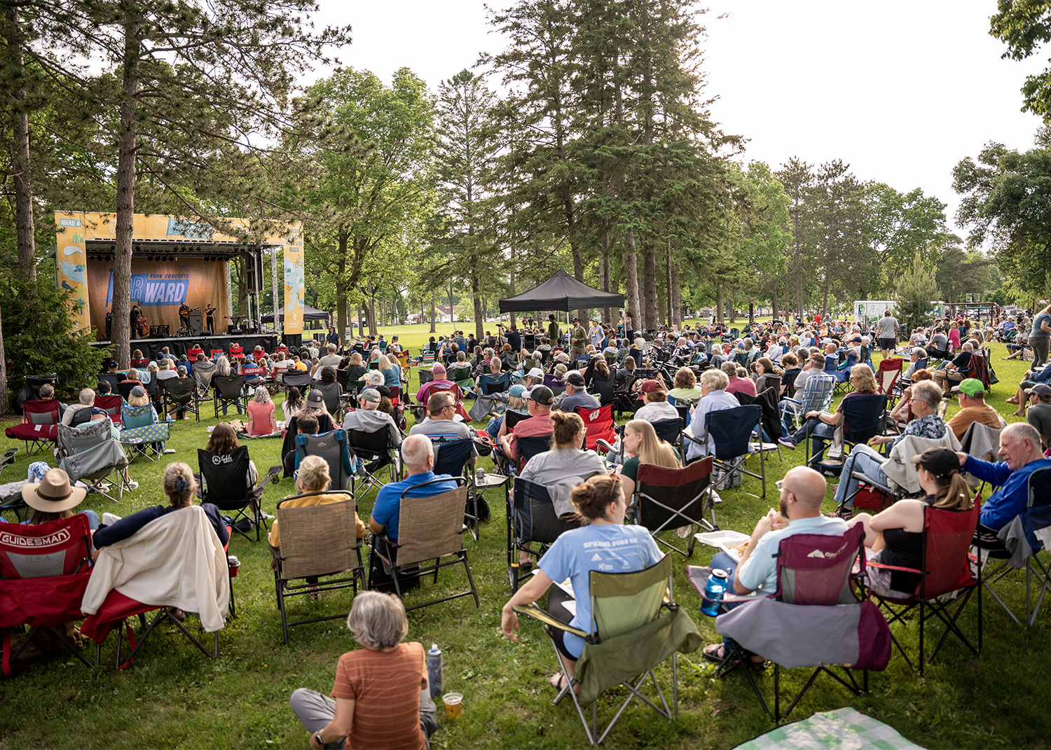 Audience seated on lawn chairs watching a live band perform on an outdoor stage at a park concert